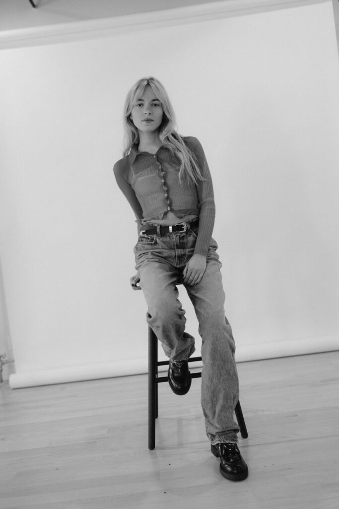 A black and white portrait of a young woman seated on a black stool, featuring a casual yet stylish outfit with a ribbed, button-up top and wide-legged jeans, complemented by chunky black boots.