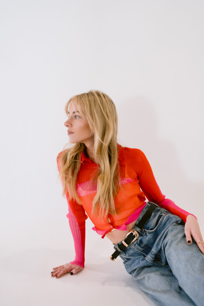 A young woman in a vibrant red and pink mesh top paired with relaxed-fit denim jeans sits casually against a white backdrop, conveying a chic and effortless style.