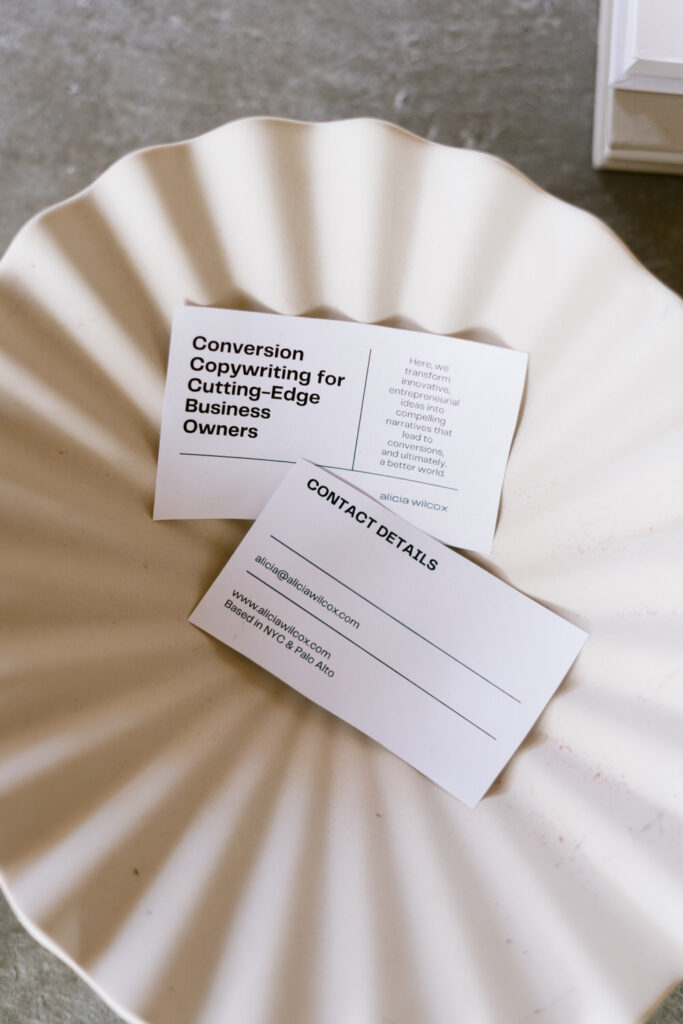 A close-up of two business cards resting on a ribbed ceramic dish, one card reading 'Conversion Copywriting for Cutting-Edge Business Owners' and the other with contact details for 'Alicia Wilcox.