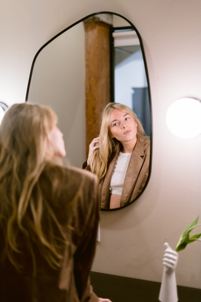 A woman in a contemplative moment, looking at her reflection in an oval mirror, with a simple white top and brown blazer complementing the warm, natural tones of the room.