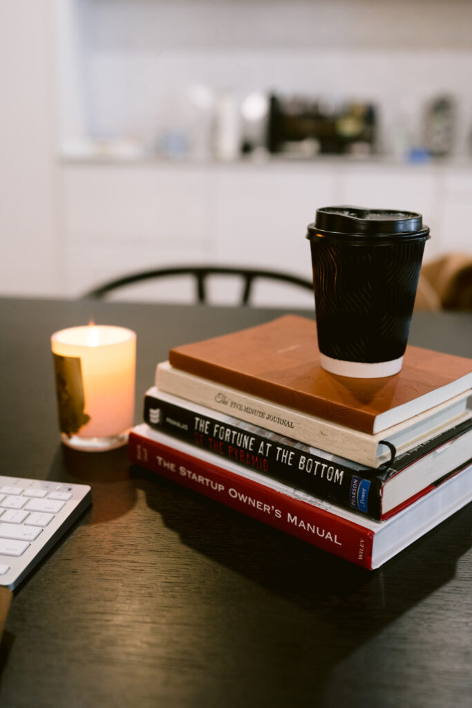 A cozy work setting featuring a stack of books including 'The Startup Owner's Manual' and 'The Fortune at the Bottom of the Pyramid', a lit candle, and a takeaway coffee cup, suggesting a peaceful reading or study session.