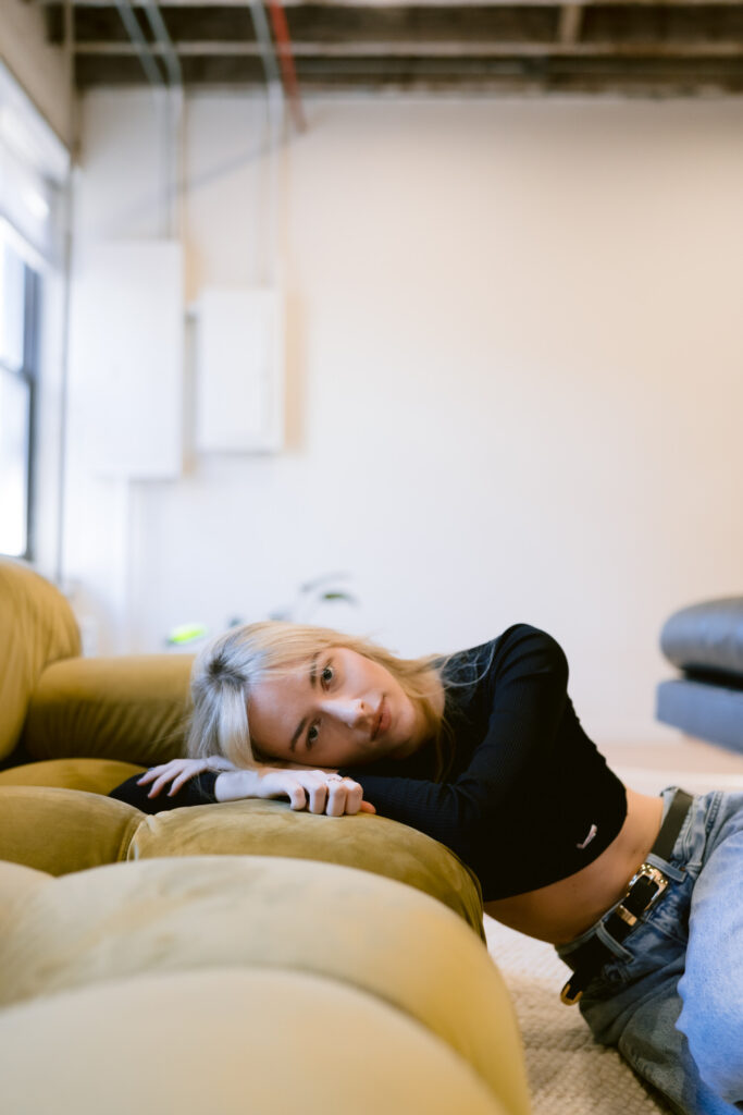 A young woman lays across a mustard couch with a contemplative expression and an industrial-chic backdrop softening the mood.