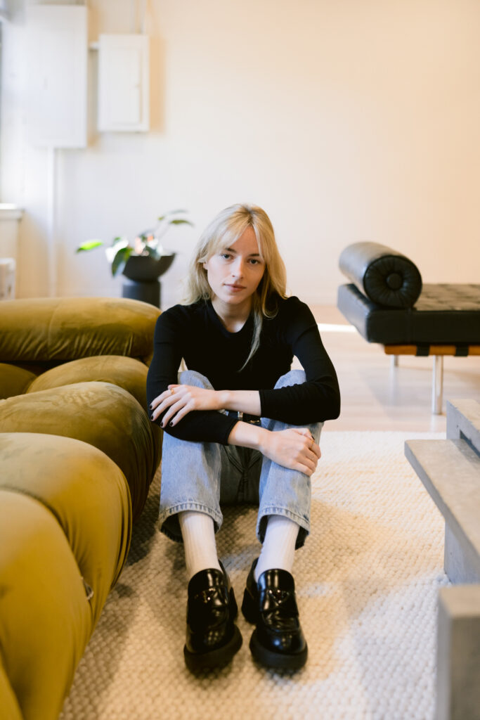 Seated on the floor, a young woman in a black top and blue jeans looks pensively towards the camera, surrounded by a cozy, modern living room with a touch of greenery.