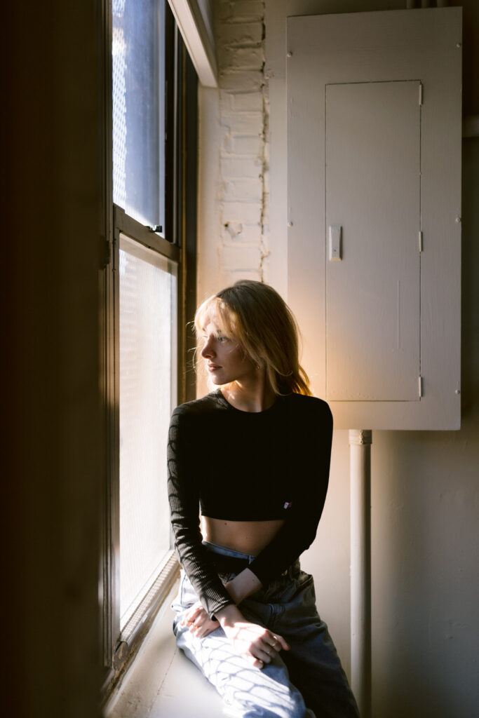A contemplative young woman gazes out a window, bathed in the gentle light of the sun, with a black cropped top and relaxed jeans.