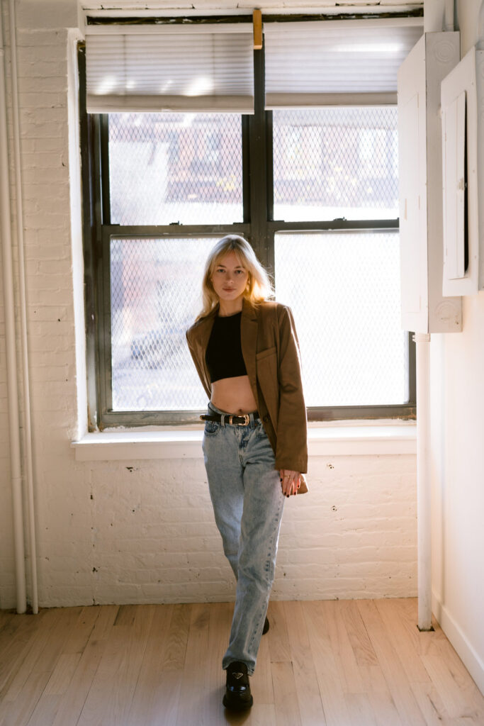 A woman stands confidently in a naturally lit room by a window, dressed in a stylish combination of a cropped top, high-waisted jeans, and a brown blazer.