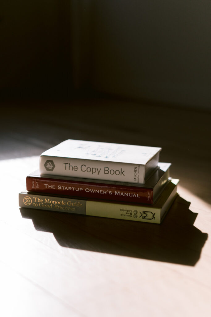A stack of books is illuminated by a shaft of sunlight on a wooden surface, featuring 'The Copy Book,' 'The Startup Owner’s Manual,' and 'The Monocle Guide to Good Business,' hinting at a theme of personal development.