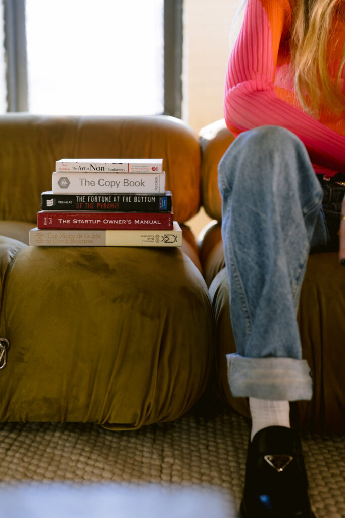 A woman sitting on a brown sofa is partially obscured by a stack of books she holds, including 'The Art of Non-Conformity' and 'The Monocle Guide to Good Business', indicating a theme of innovative thinking and entrepreneurship.