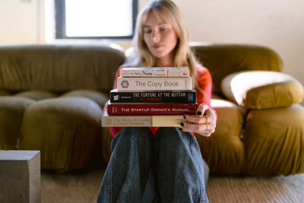 A woman sits in a cozy room holding a stack of books in front of her face, including titles like 'The Art of Non-Conformity,' 'The Copy Book,' 'The Fortune at the Bottom of the Pyramid,' 'The Startup Owner’s Manual,' and 'The Monocle Guide to Good Business,' indicating a theme of entrepreneurial and creative inspiration.