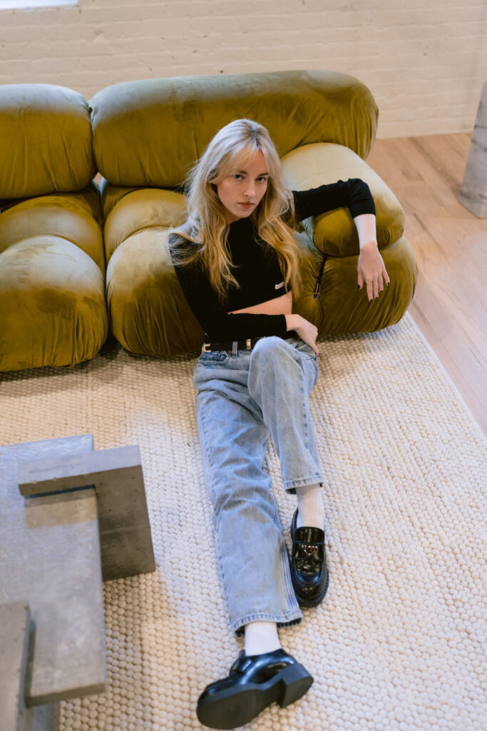 A casual yet assertive pose by a woman seated on a plush green sofa, clad in a fitted black top and blue jeans, with stark modern decor contrasting the warm brick background of an urban loft space.
