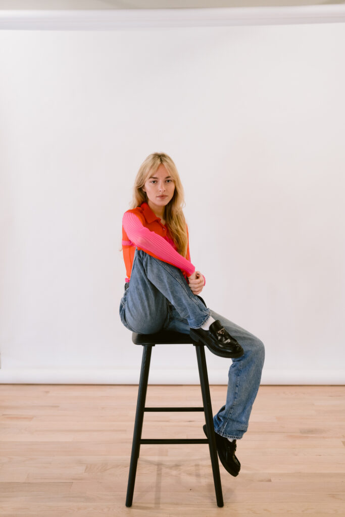 Seated on a tall black stool, a young woman poses with her knee hugged to her chest, wearing a bold orange and pink top paired with classic blue jeans, set against a simple white backdrop for a minimalistic style statement.