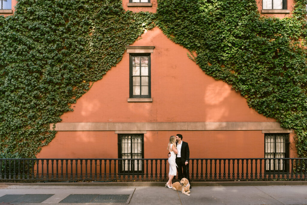 A couple hugs one another with their dog against a brick building covered in greenery.