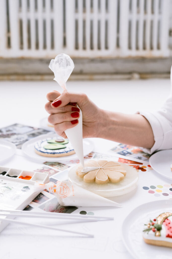 A hand with red nail polish decorates a flower-shaped cookie with white icing using a piping bag, surrounded by other decorated cookies and art supplies on a white table.