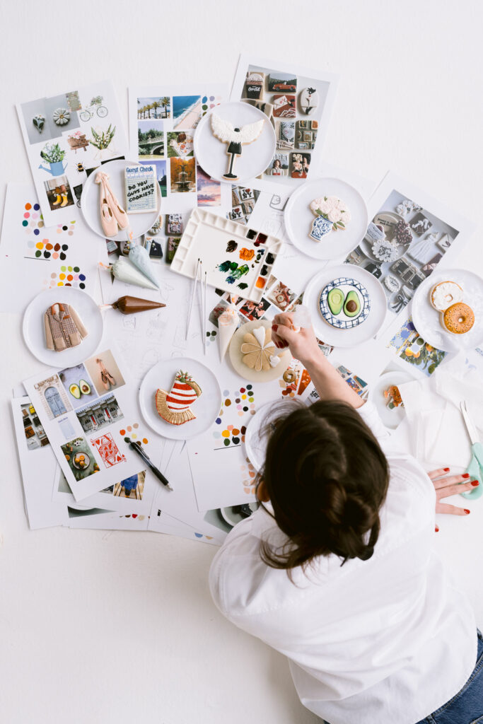 An overhead view of a woman in a white shirt decorating cookies on a table covered with art supplies, color palettes, and photos of decorated cookies.