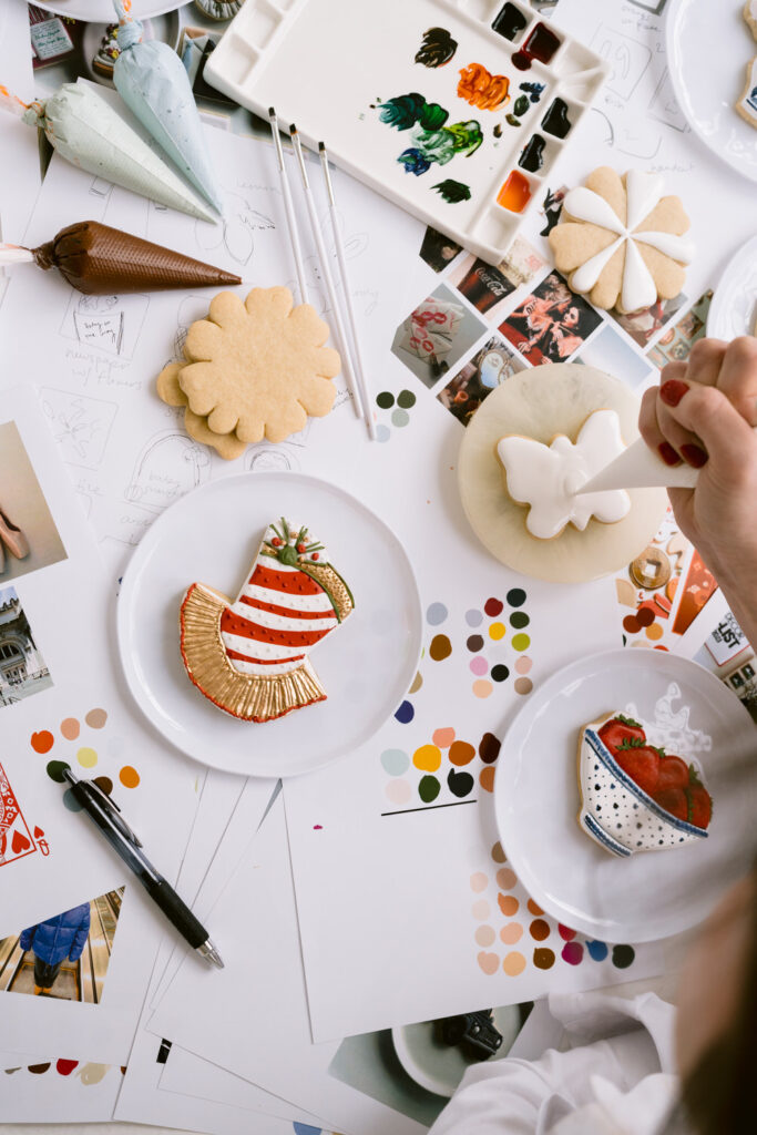 Close-up of a hand with red nail polish using a piping bag to decorate a butterfly-shaped cookie with white icing, surrounded by other decorated cookies, color palettes, and art supplies on a white table.