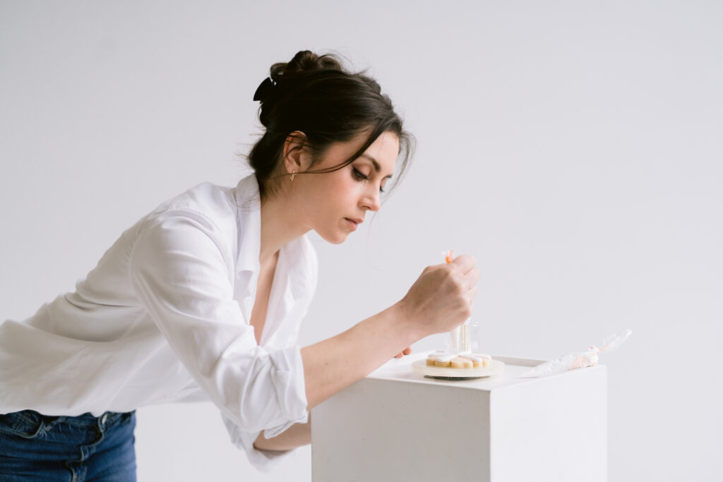 A woman with dark hair in an updo decorates a cookie using a piping bag, leaning over a white block and focused on her work, wearing a white button-down shirt and blue jeans.