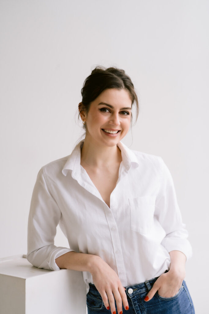 A woman with dark hair styled in an updo smiles while leaning on a white block, wearing a white button-down shirt and blue jeans.