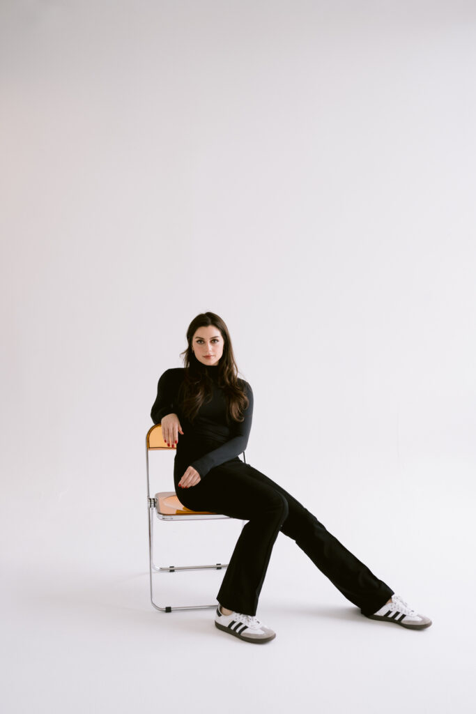 A woman with long dark hair wearing a black turtleneck sits on a chair with her legs extended, leaning slightly back, against a plain white background.