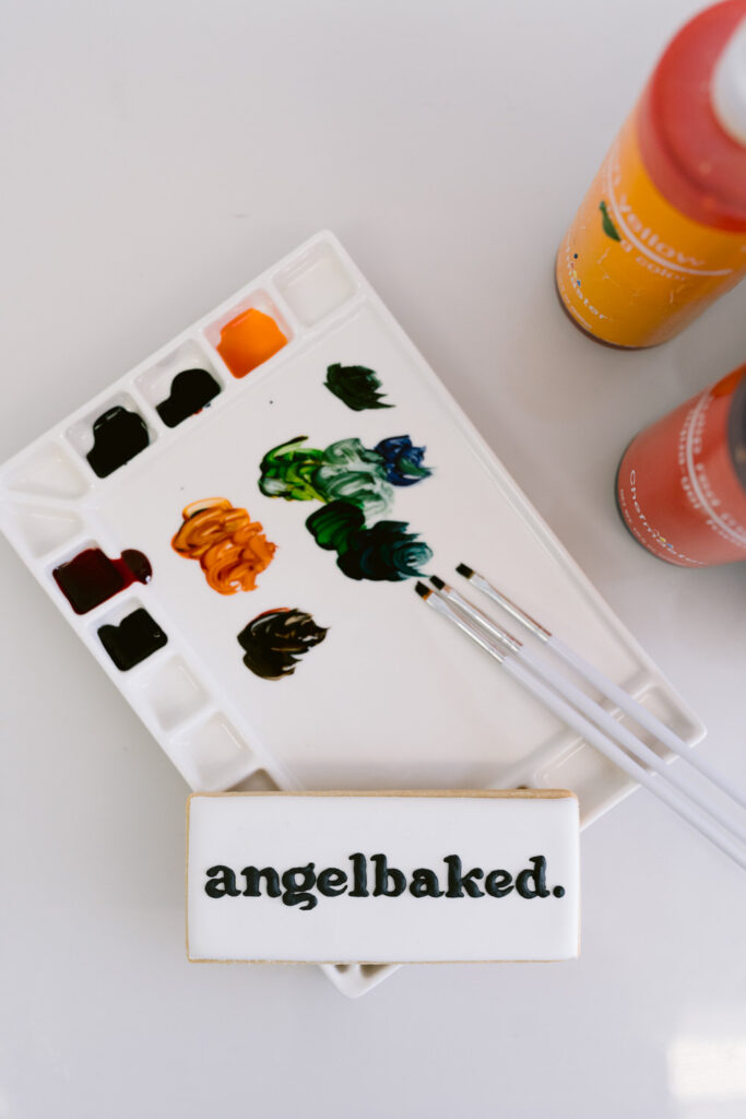 A white rectangular cookie with the text 'angelbaked.' placed next to a paint palette with colorful paints and brushes, along with orange and yellow bottles on the side.