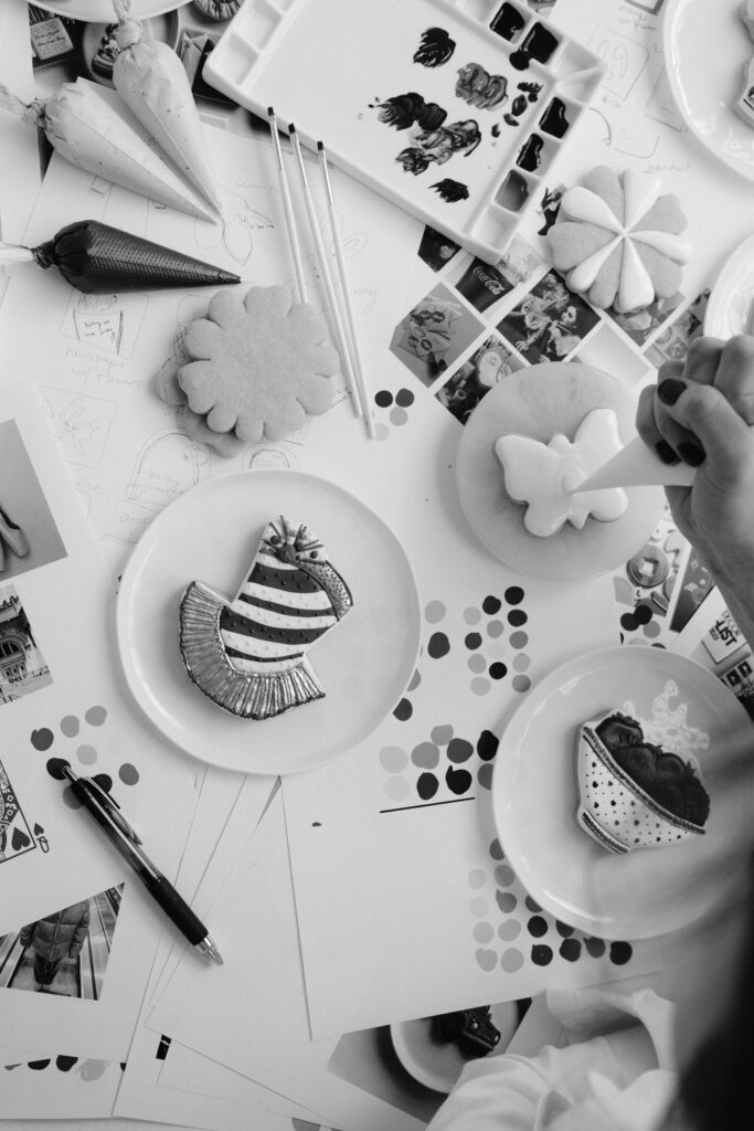 Black and white image of a hand decorating a butterfly-shaped cookie with white icing, surrounded by other decorated cookies, color palettes, and art supplies on a white table.