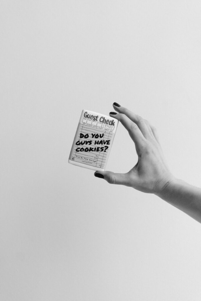 Black and white image of a hand holding a rectangular cookie decorated like a guest check that says, 'Do you guys have cookies?' against a plain background.