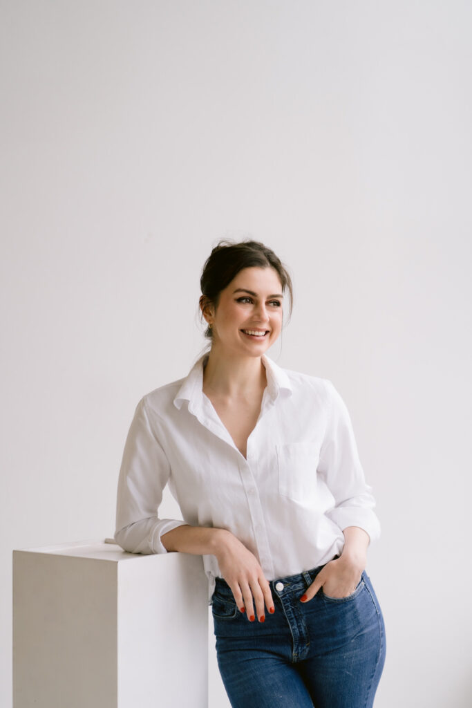 A woman with dark hair styled in an updo smiles while leaning on a white block, wearing a white button-down shirt and blue jeans.