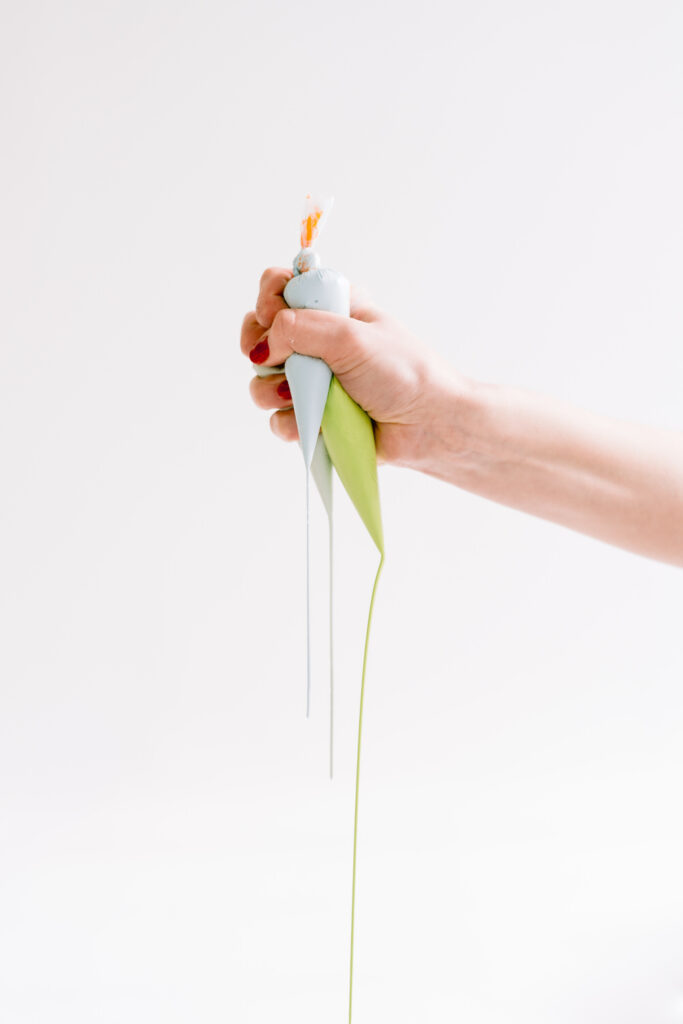 A hand with red nail polish squeezes two piping bags filled with light blue and green icing, with the icing streaming out, against a plain white background.