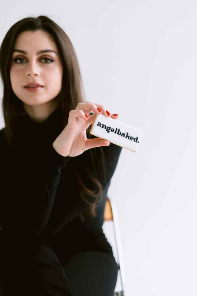 A woman with long dark hair wearing a black turtleneck holds a rectangular cookie with the text 'angelbaked.' on it.