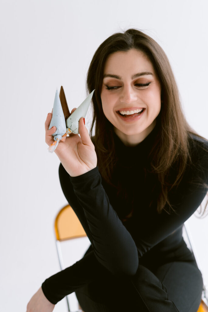 A woman with long dark hair smiles while holding two piping bags filled with different colors of icing, wearing a black turtleneck and sitting on a chair.