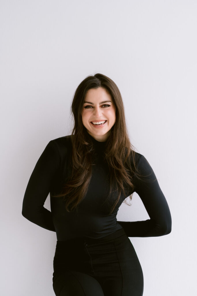 A woman with long dark hair smiles while wearing a black turtleneck, standing against a white background.