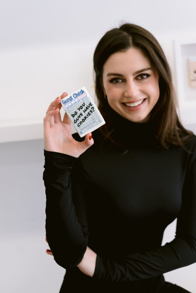 A woman with long dark hair smiles while holding a rectangular cookie decorated like a guest check that says, 'Do you guys have cookies?', wearing a black turtleneck.