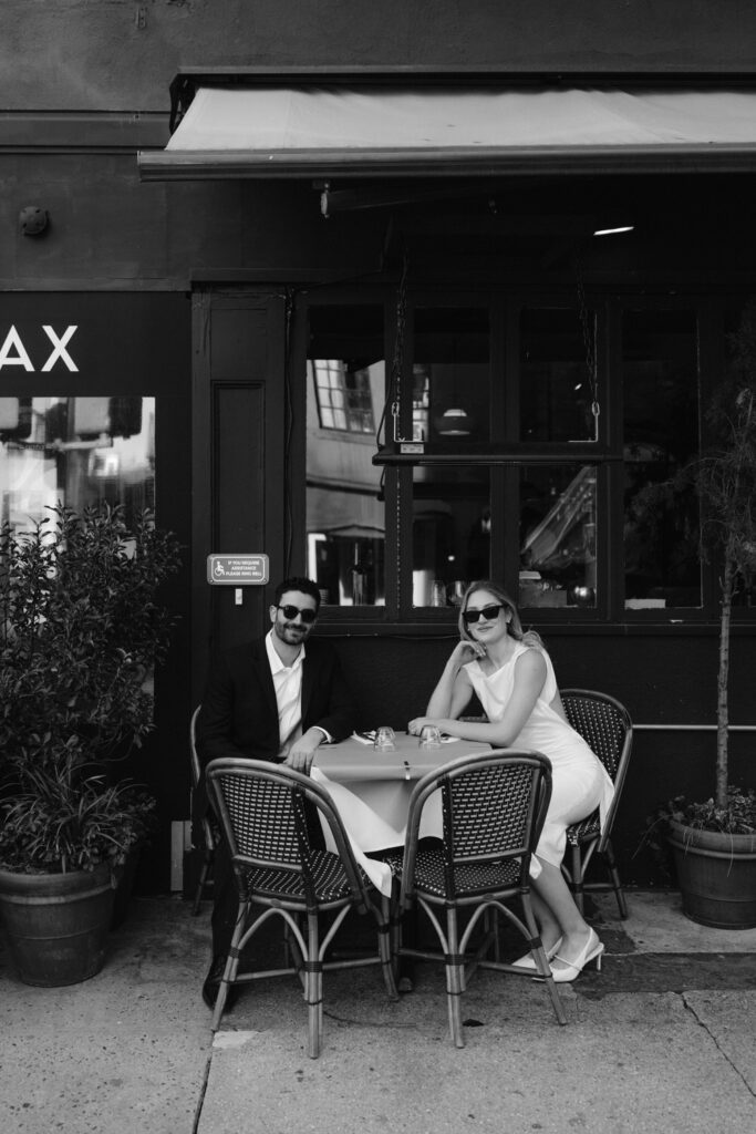 A couple sits at a table outdoors with sunglasses on.
