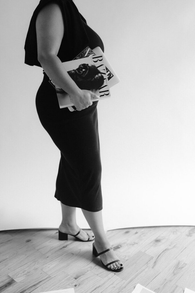 Black and white photo of a person holding a stack of magazines by her side. 
