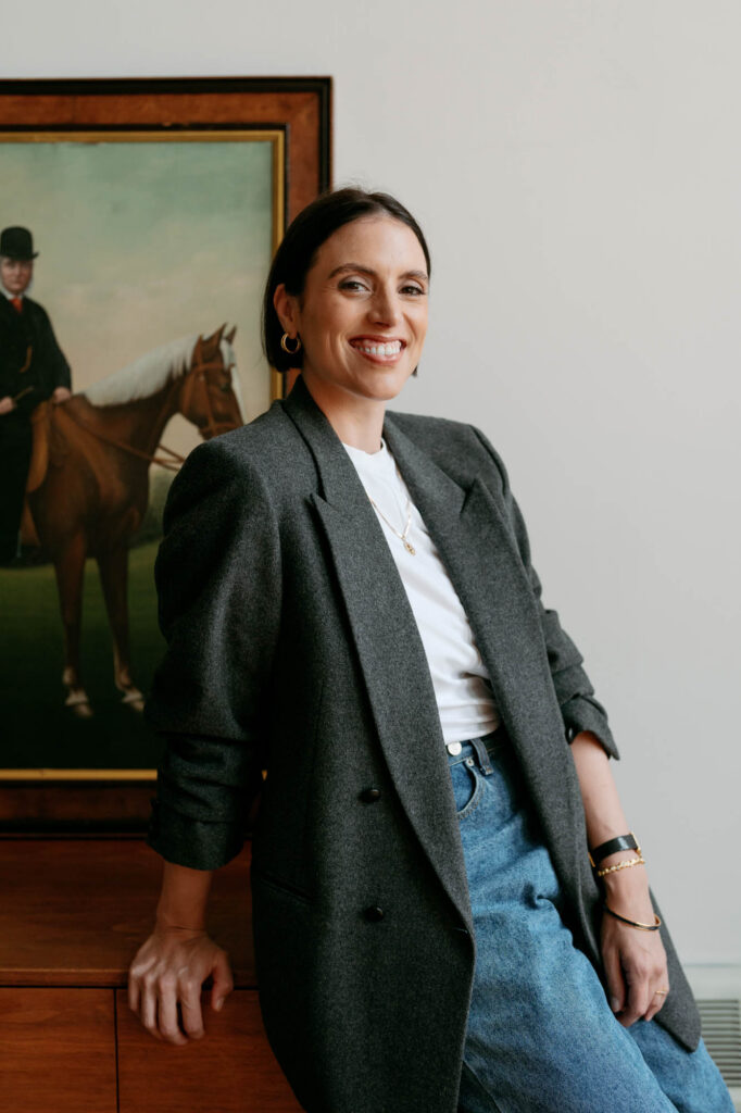 Zay smiling in a grey blazer and white tee, leaning on a walnut credenza beside a vintage equestrian painting