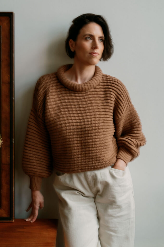 Zay in a camel chunky knit sweater and cream jeans leaning against a white wall, looking toward the light