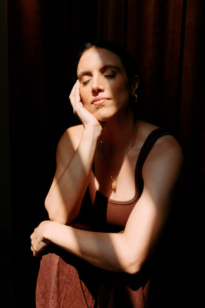 Zay with eyes closed in a beam of direct window light, wearing a brown tank and gold pendant necklace