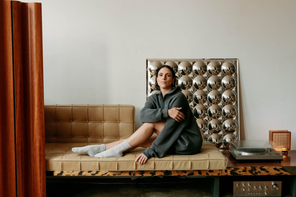 Pilates instructor Zay in a grey sweatshirt dress seated on a tan leather daybed during her Brooklyn personal branding photoshoot