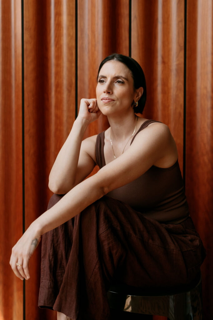 Zay gazing toward the window in a brown tank against warm wood paneling in a Brooklyn studio